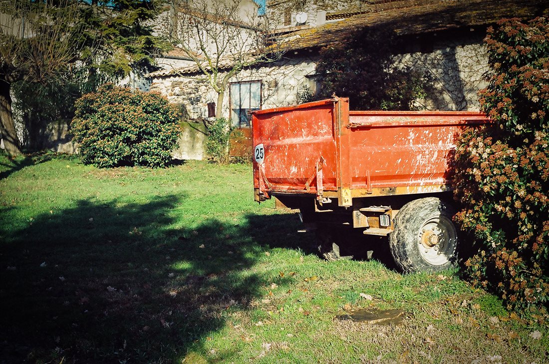 Ardèche, partie arrière d'une maison d'agriculteur, herbe verte, bâtiments en pierres en fond, une ombre sur le sol, lumière vive et chaude, arrière d'une remorque de tracteur orangée à moitié cachée par la végétation